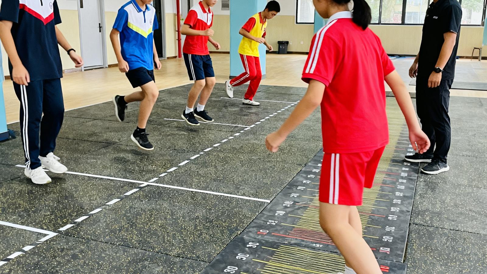 zoty体育分校区内，6名初三学生在专业教练的指导下分组进行立定跳远技术动作分解练习，地面铺设防滑塑胶垫并标有刻度线。